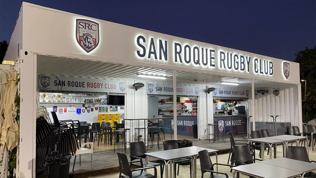 Outdoor seating area in front of San Roque Rugby Club, a white building with illuminated signage and club logos, photographed at dusk.